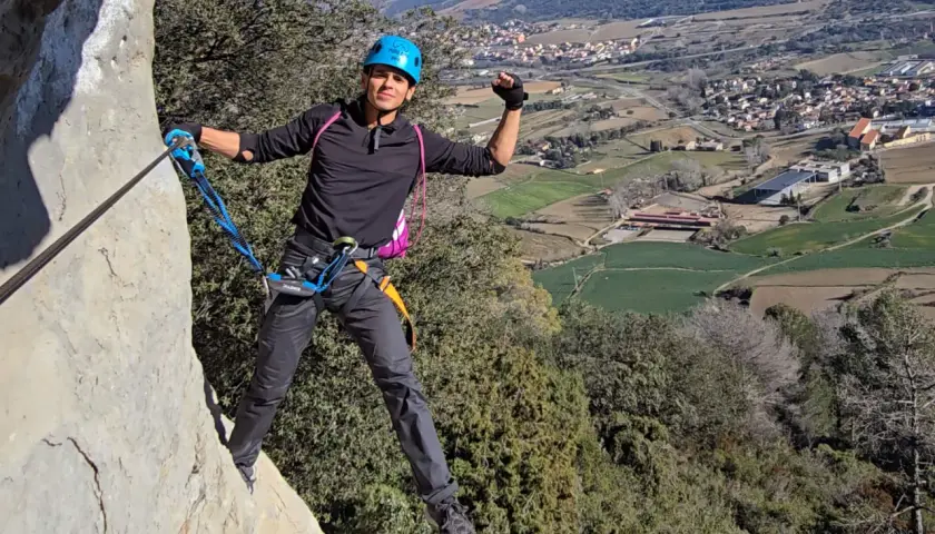 Un escalador sonriente, con casco azul, disfruta de la vía ferrata, un ejemplo de los diversos tipos de escalada, con un pueblo y campos verdes de fondo.
