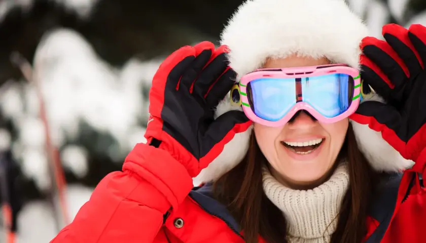 Mujer sonriente con gorro de piel y gafas de esquí rosas, lista para la nieve. Explora las categorías de gafas de esquí para encontrar tu estilo.