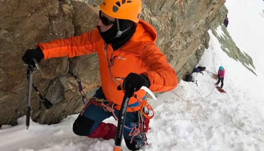 Alpinista con casco naranja y chaqueta brillante usa un piolet para escalar una pared rocosa nevada.
