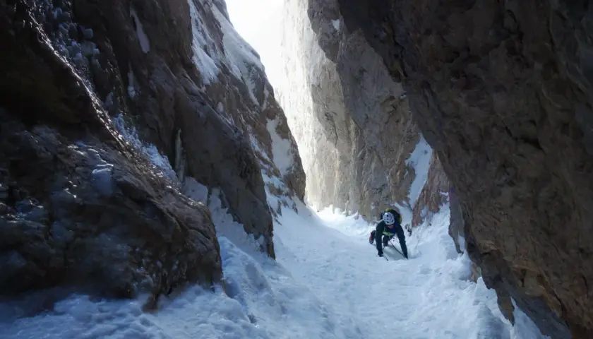 Alpinista asciende por un estrecho cañón nevado, usando piolet y crampones. La estaca de nieve se ve firme bajo sus pies.