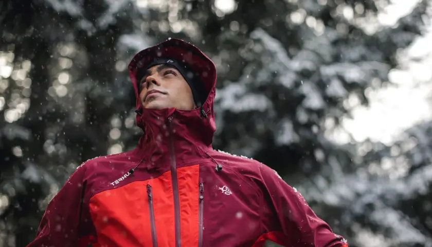 Hombre con ropa para ir a la montaña, con capucha y chaqueta impermeable, bajo una nevada.