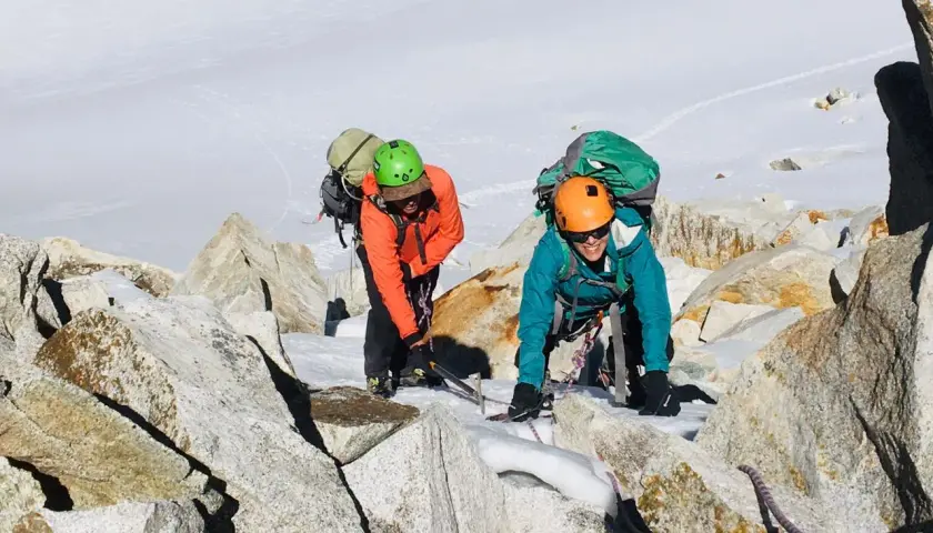Dos escaladores con mochilas y cascos navegan por rocas y nieve en la montaña. Sus aventuras son como libros abiertos de la naturaleza.
