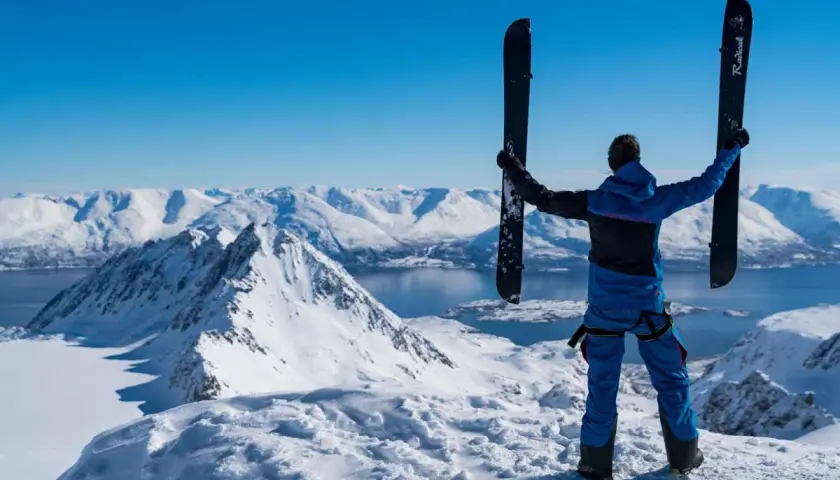 Persona en la cima de una montaña nevada, sosteniendo un splitboard que es un equipo versátil para ascender y descender.