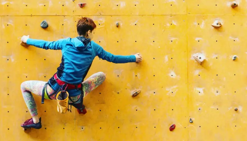 Una escaladora se enfrenta a una pared amarilla, buscando el siguiente agarre en una escalada de dificultad.