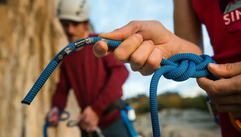 Manos atando nudos de escalada con una cuerda azul. En el fondo, otro escalador con casco.
