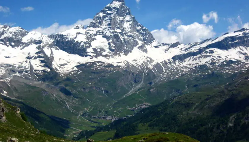 Majestuoso pico nevado se alza sobre un valle verde con un pueblo. El camino de senderismo gr5 etapes serpentea entre las colinas.