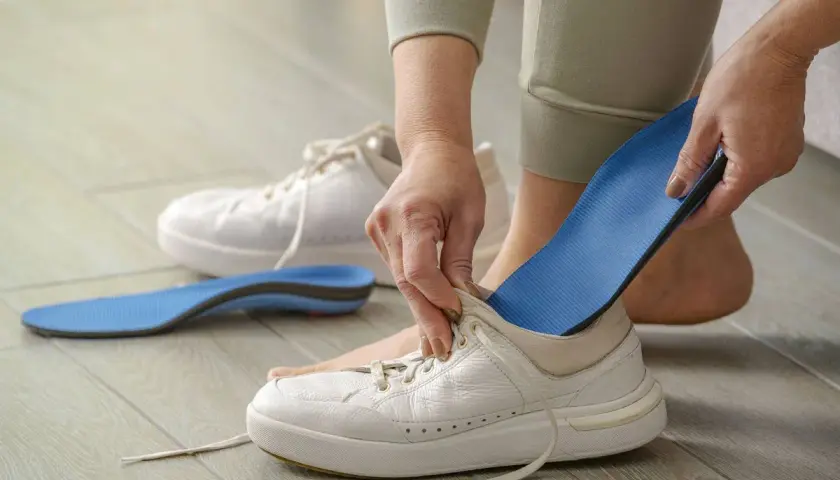 Manos insertando una plantilla azul en una bota blanca. Otra plantilla y una bota están en el suelo.