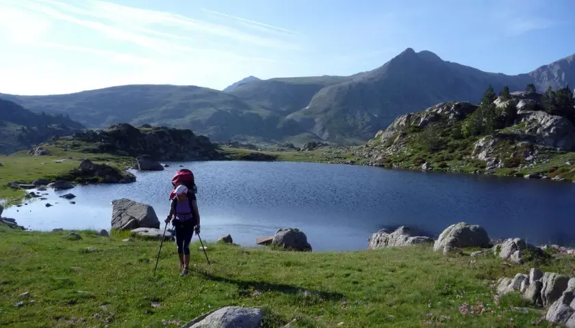 Senderista con mochila y bastones camina por un prado verde hacia un lago azul en los Pirineos de GRP Andorra.