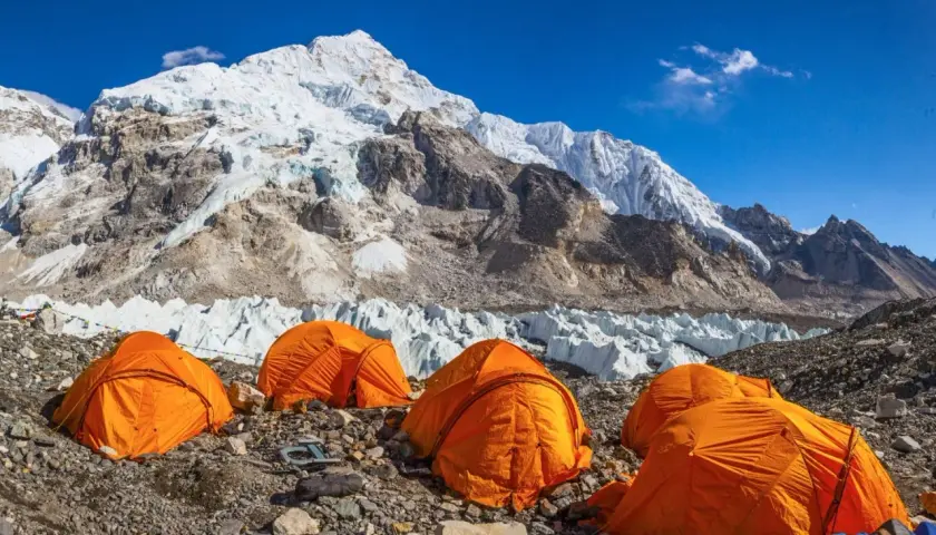 Campamento de expedición en la base de la cordillera más alta del mundo, con tiendas naranjas y glaciares imponentes bajo un cielo azul.
