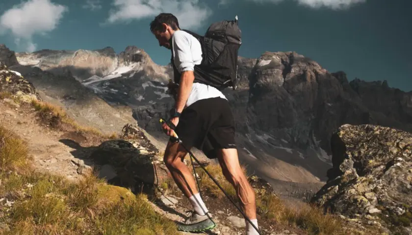 Kilian Jornet asciende una ladera rocosa en los Pirineos, con bastones y mochila, bajo un cielo azul con nubes.