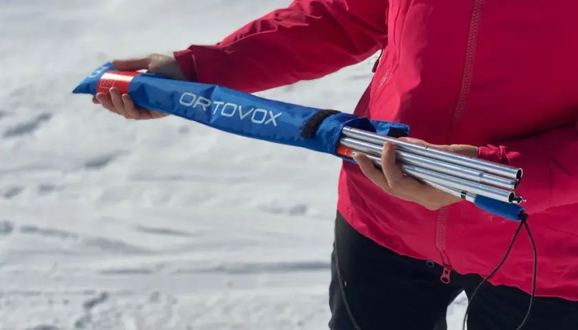 Persona con chaqueta rosa sostiene una arva, sonda y pala ORTOVOX en una bolsa azul, lista para la aventura en la nieve.