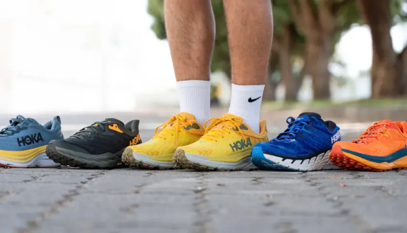 Cinco pares de zapatillas Hoka con gran amortiguación, en colores azul, gris, amarillo, azul y naranja, alineados en un camino.