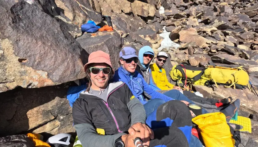 Cuatro montañistas sonríen en la ladera rocosa, listos para la aventura. El **bru busom** de uno de ellos, un sombrero rosa, destaca entre el equipo de expedición.