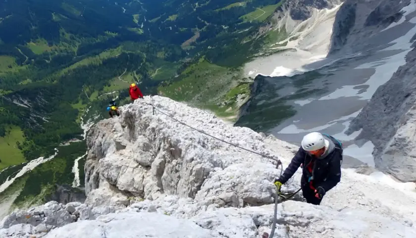 Escaladores en una vía ferrata, asegurados a un cable de acero que recorre la cresta rocosa. El paisaje montañoso es espectacular.