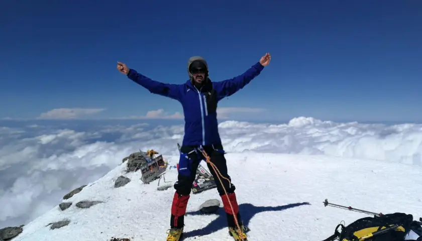 Javier García Díaz, alpinista, celebra en la cima nevada, rodeado de nubes y un cielo azul intenso.