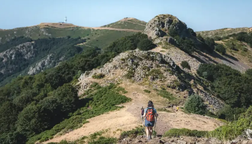 Senderismo en montaña con bastones, comparando rutas en AllTrails vs. Wikiloc. Paisaje verde y rocoso bajo un cielo azul.
