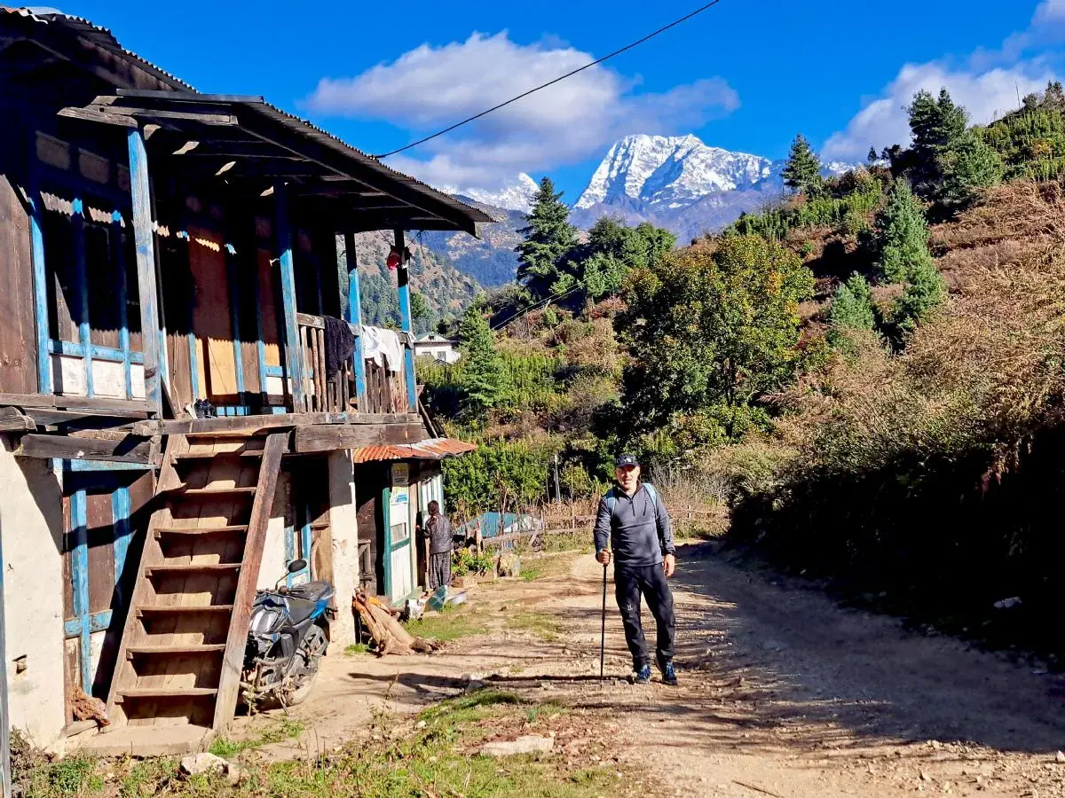 Hombre con bastón de trekking en un camino rural, con una casa tradicional y montañas nevadas al fondo. Una aventura de trekking Himalaya.