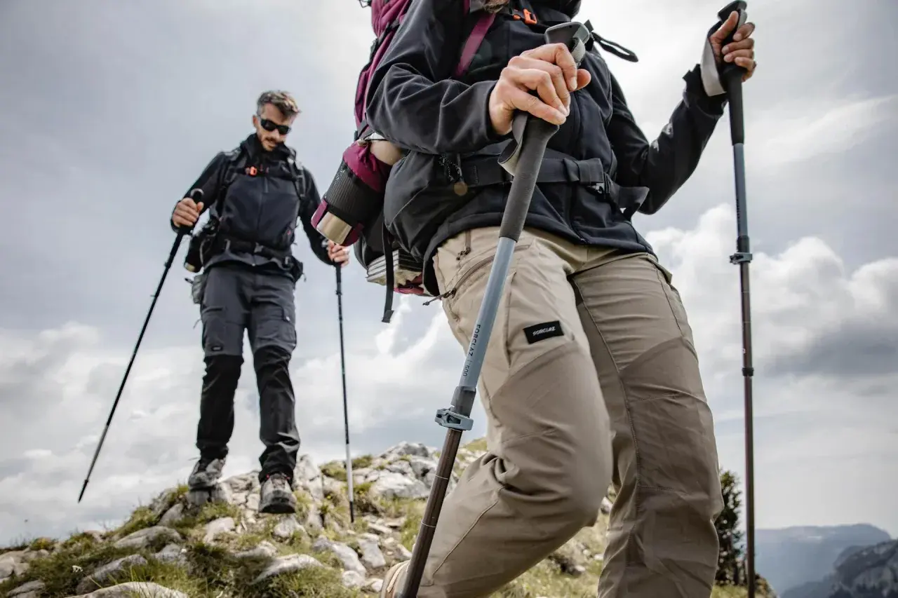 Dos excursionistas suben una ladera rocosa, usando sus pals per caminar para mayor estabilidad. El cielo nublado sugiere una aventura desafiante.