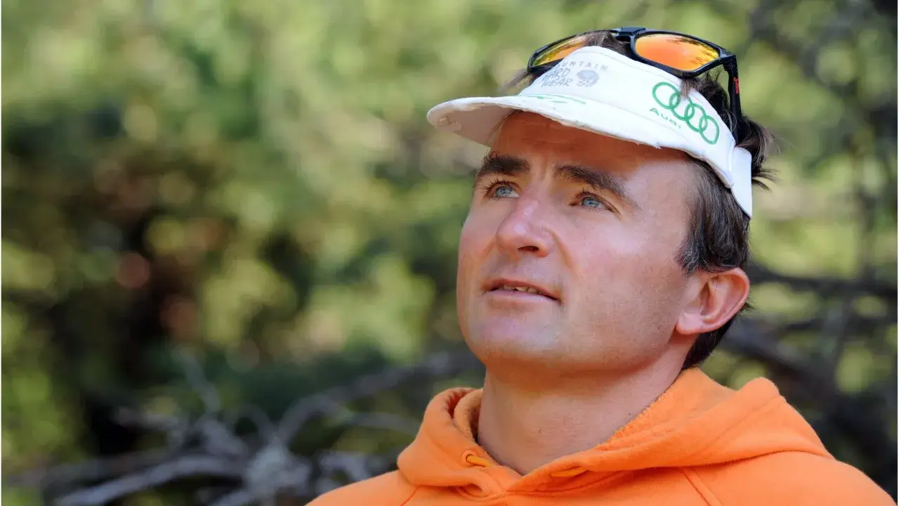 Ueli Steck, con gorra blanca y sudadera naranja, mira hacia arriba con determinación.
