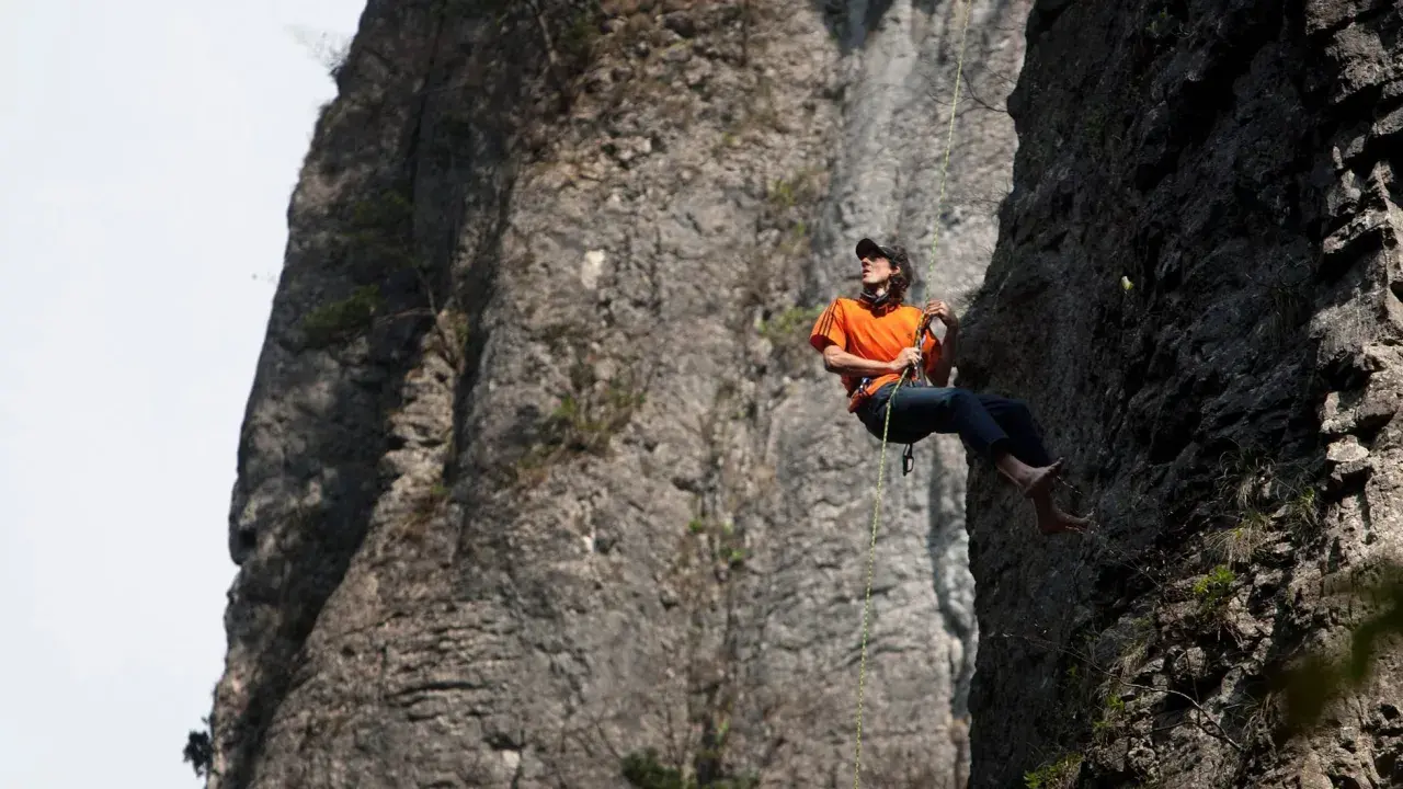 Dean Potter desciende por una pared rocosa, con el sol brillando sobre su camiseta naranja.