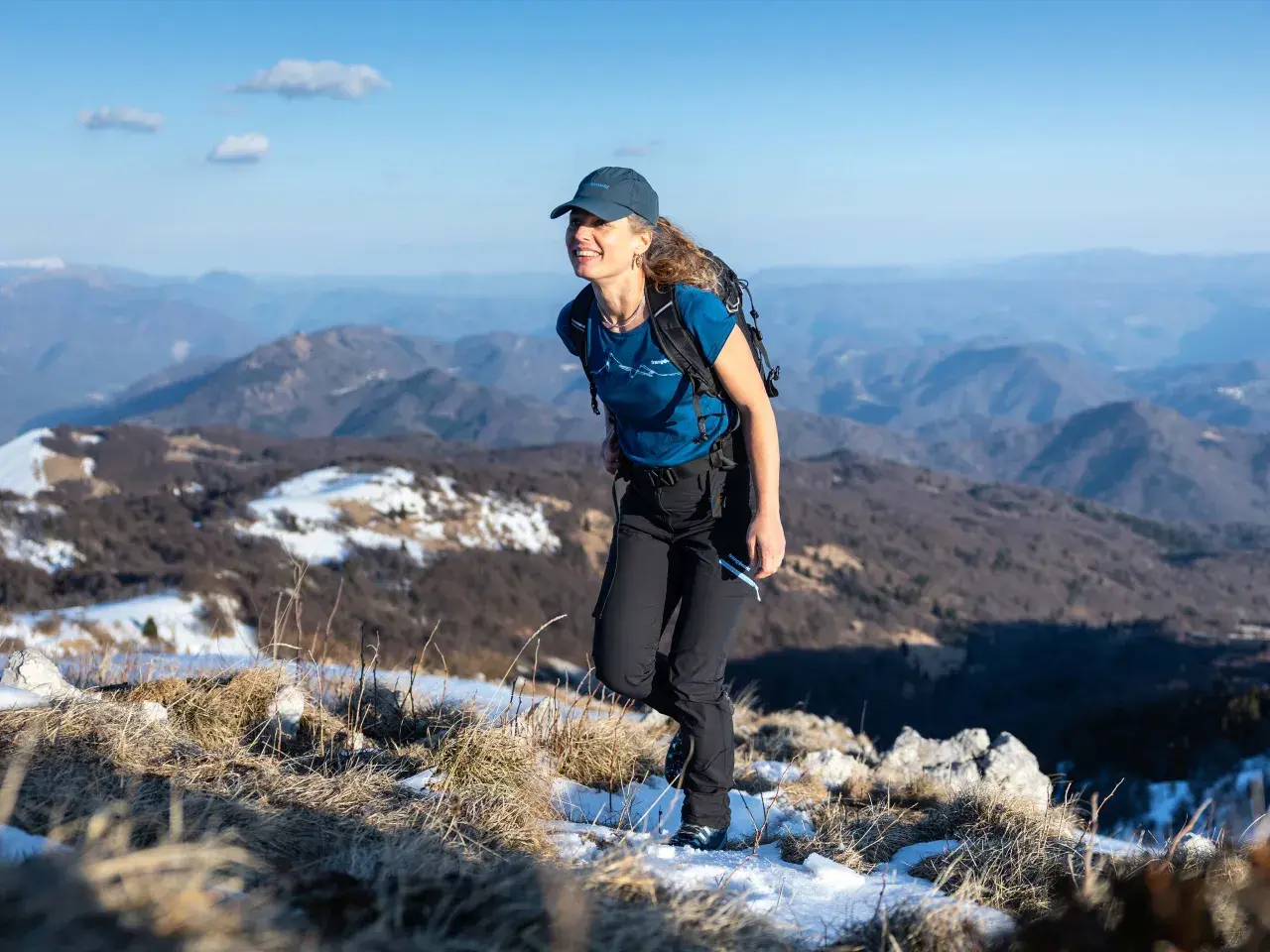 Mujer sonriente camina por la montaña con ropa de abrigo, incluyendo una camiseta de lana merino, disfrutando del paisaje nevado.