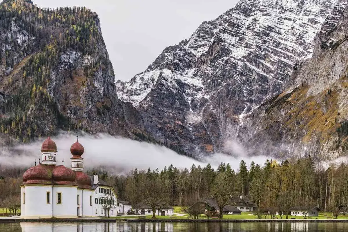 Iglesia blanca con cúpulas rojas junto a un lago, rodeada de bosques y majestuosas montañas nevadas en los Alpes alemanes.