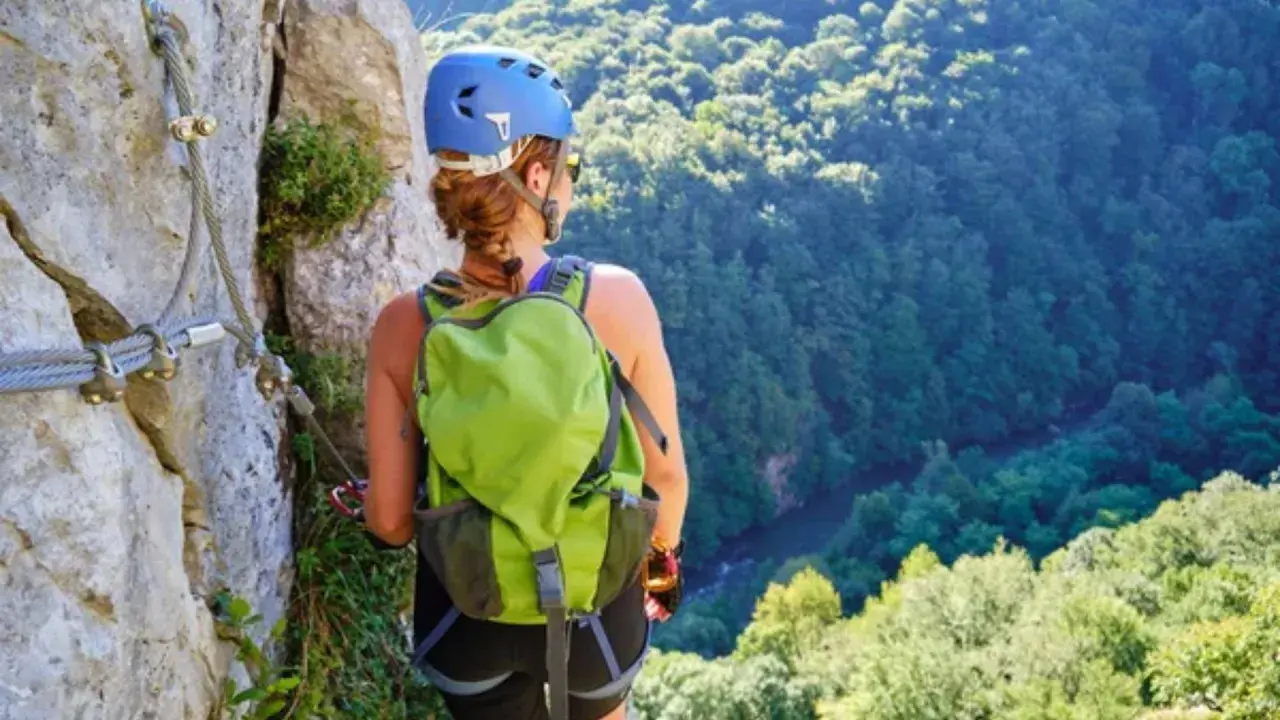 Mujer con casco y mochila verde en vía ferrata, con vistas a un bosque frondoso. Afortunadamente, no hubo ningún accidente de escalada.