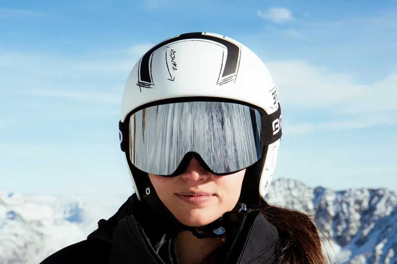 Mujer con casco de esquí blanco y gafas espejadas, lista para deslizarse por la montaña.
