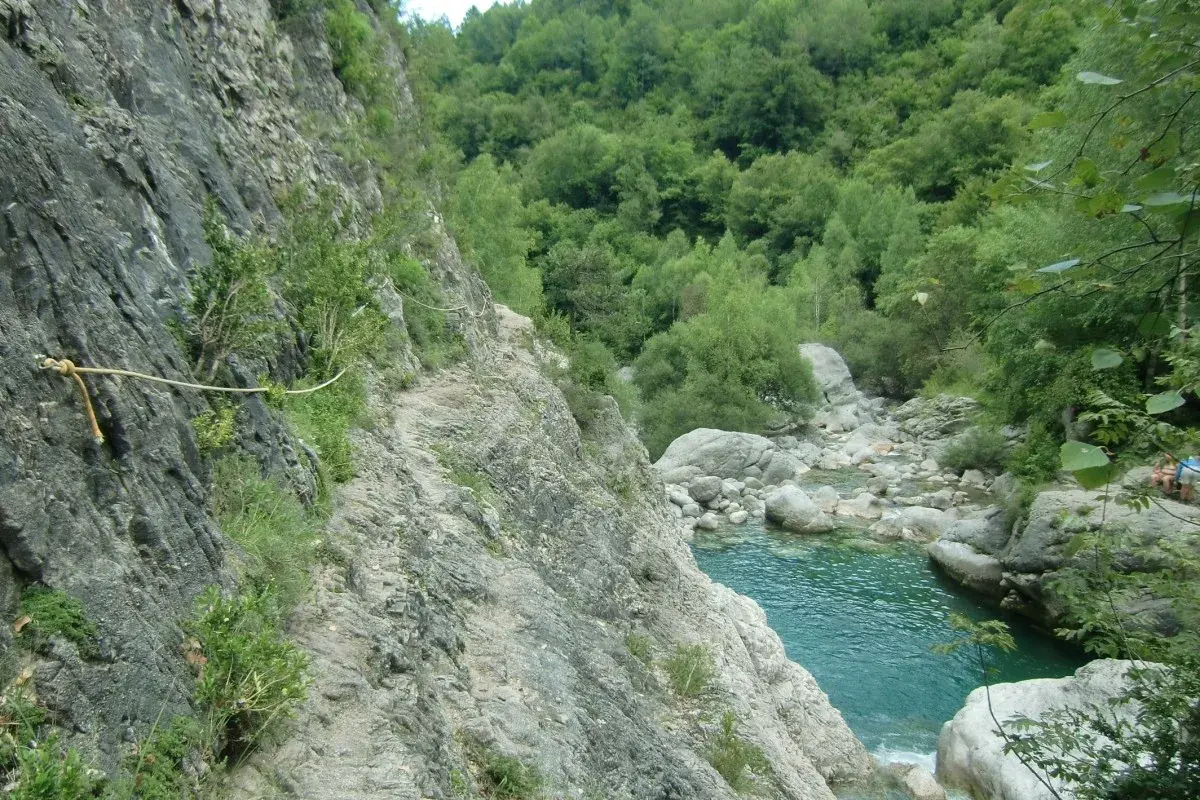 Sendero rocoso con cuerda de seguridad a lo largo de la pared de la garganta de Escuain, con un río turquesa y vegetación exuberante al fondo.