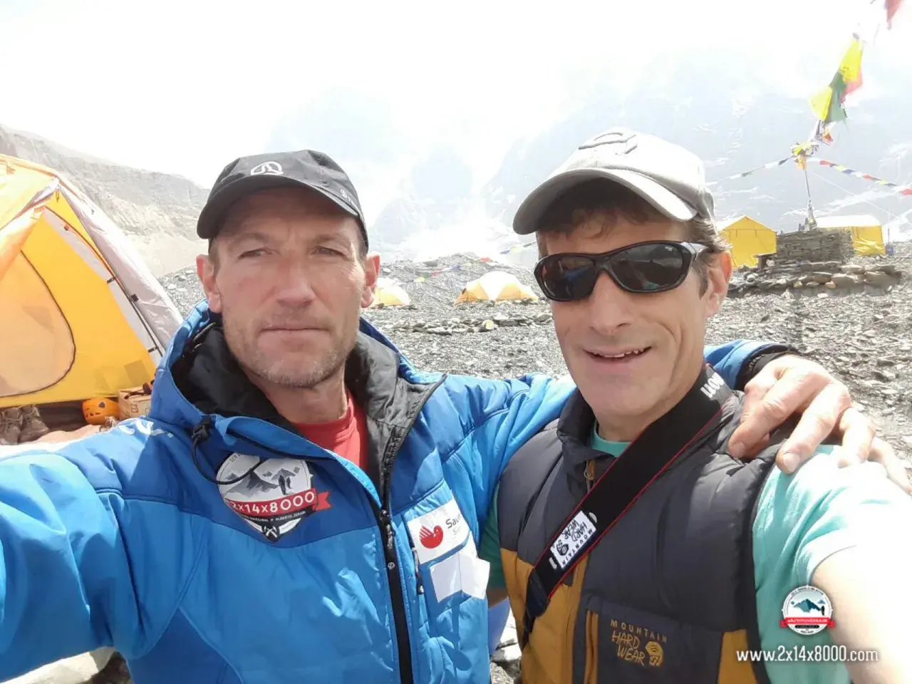 Alberto Zerain y un compañero posan sonrientes en un campamento de montaña, con tiendas amarillas y banderas de oración al fondo.
