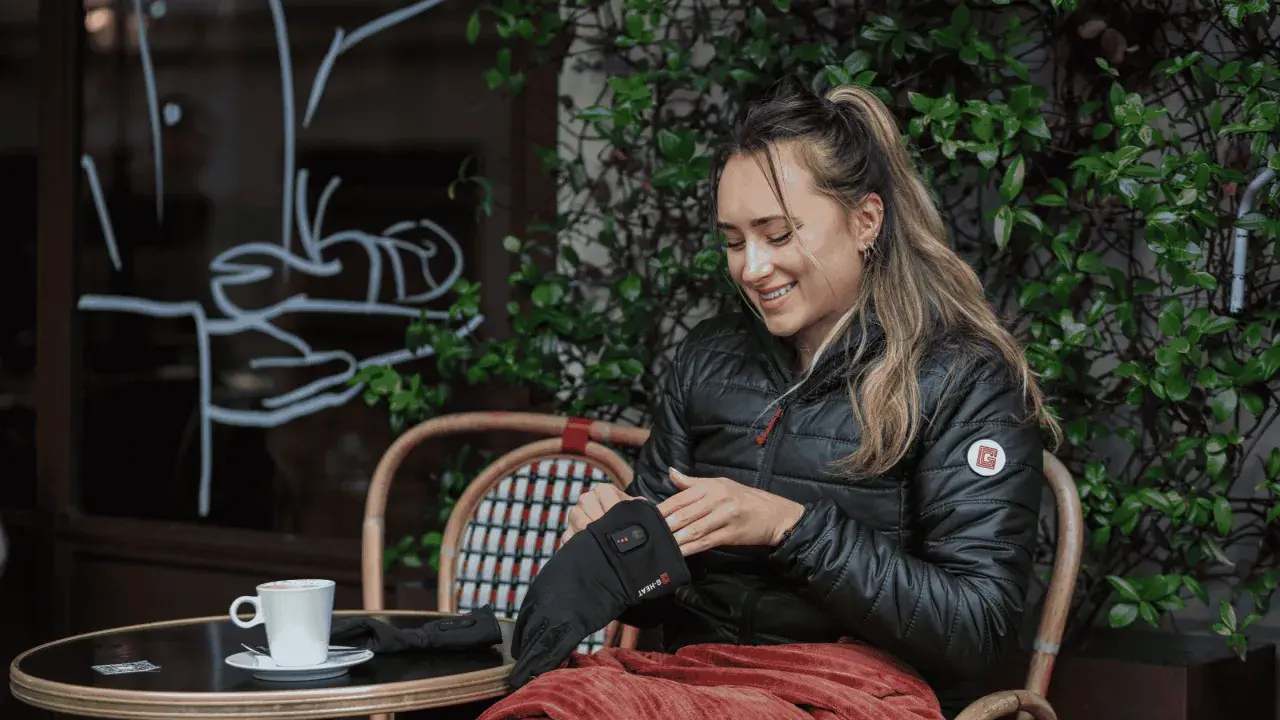 Mujer sonríe mientras se pone guantes térmicos, perfectos para el frío. Una taza de café la espera.
