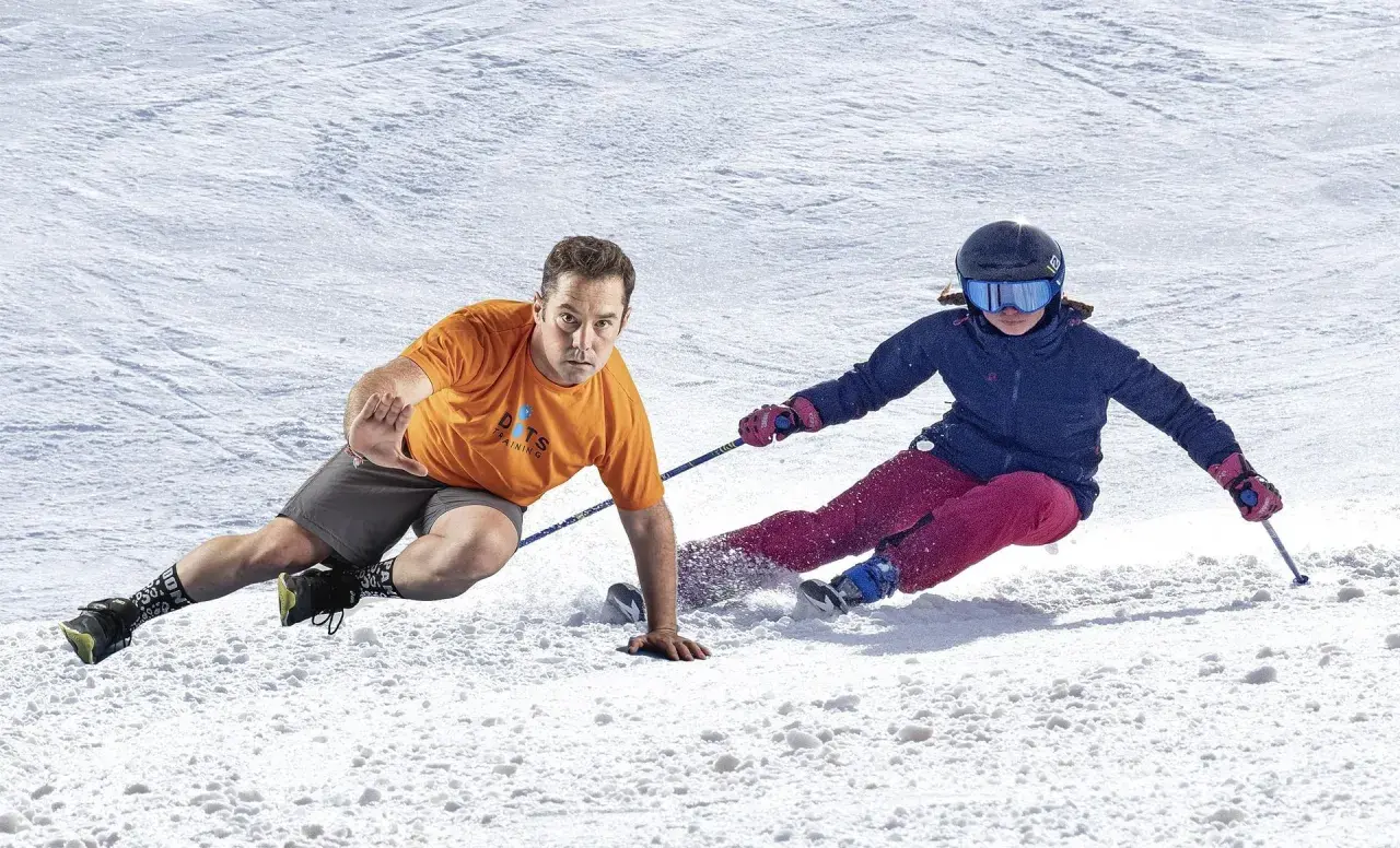 Instructor demuestra ejercicios para esquiar, con un esquiador en la nieve.