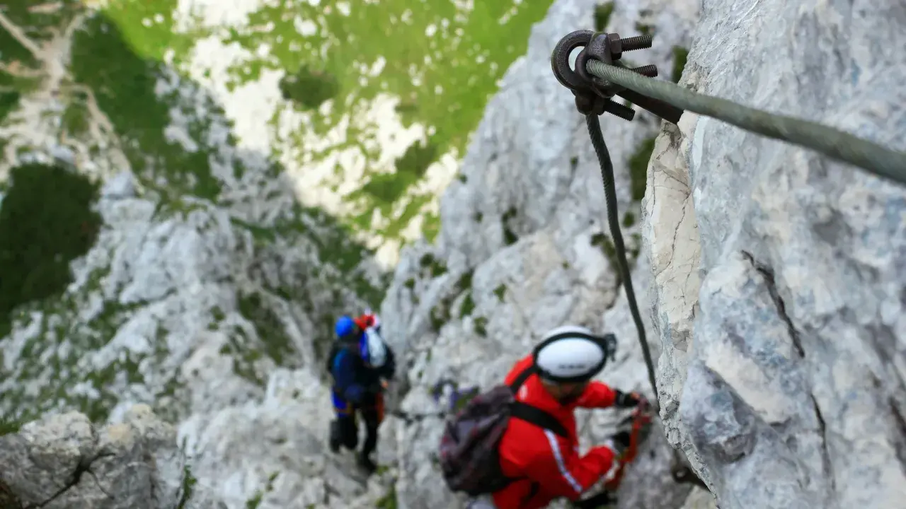 Dos personas en una vía ferrata de iniciación, aseguradas a un cable de acero mientras ascienden por una pared rocosa.