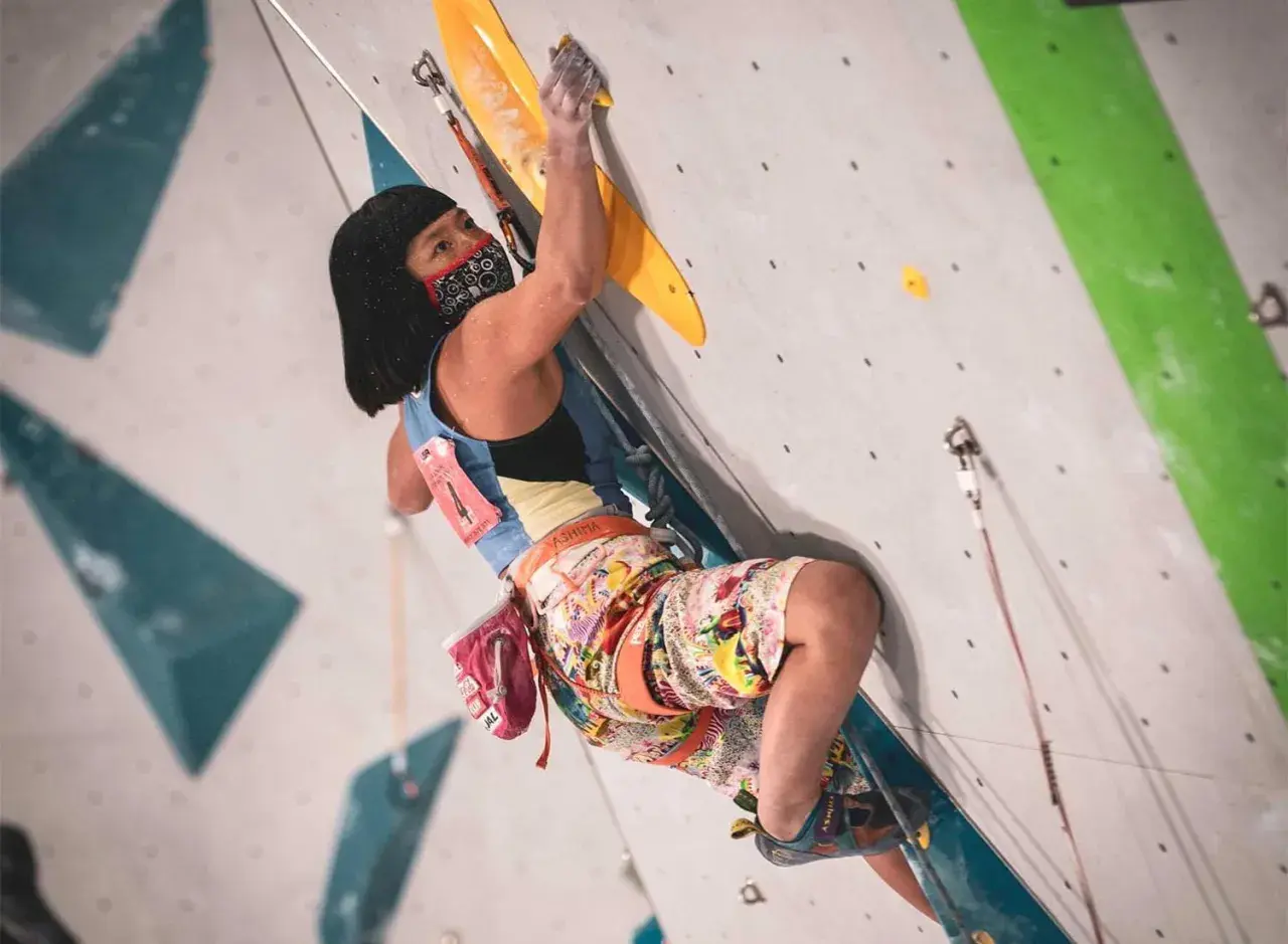 Ashima Shiraishi, con mascarilla, escala un muro de roca artificial, concentrada en cada movimiento.
