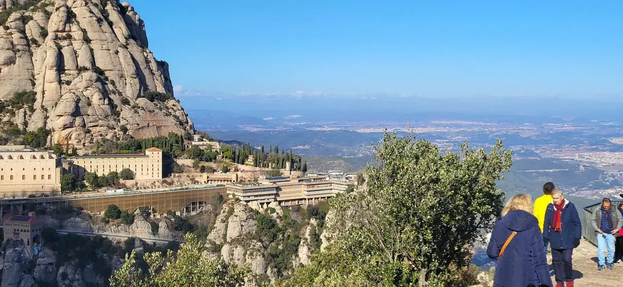 Vista panorámica del monasterio de Sant Miquel de Montserrat, con sus edificios anclados a la montaña y el valle extendiéndose hasta el horizonte.