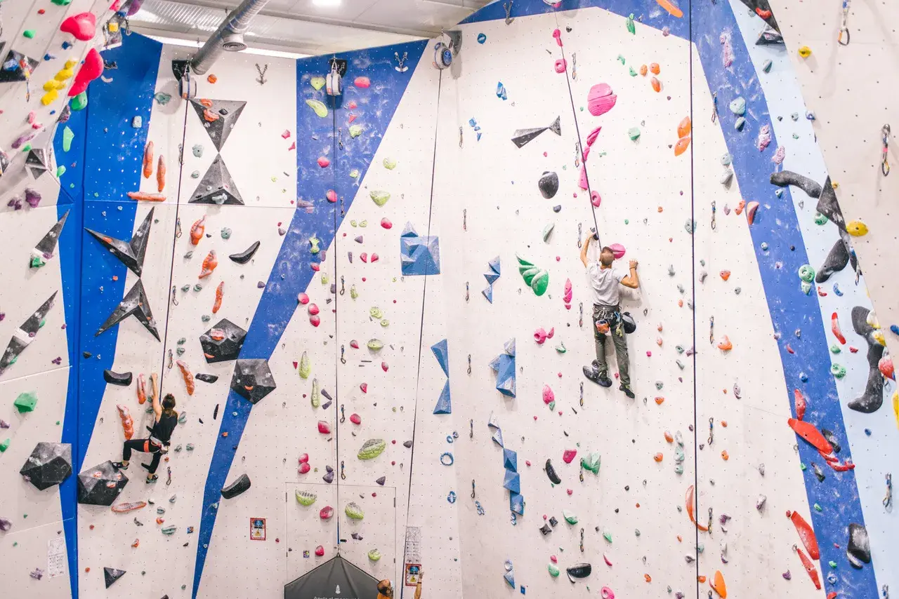 Escaladores en rocódromo, uno asciende con un asegurador automático de escalada, el otro en la pared azul.