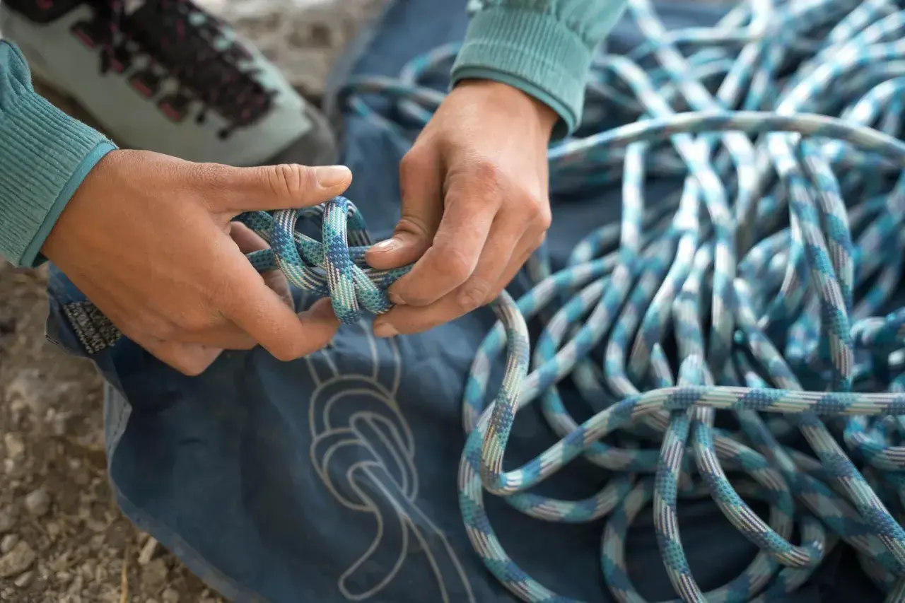 Manos preparan cuerda para machard escalada. Botas de montaña y lona azul con nudos dibujados.