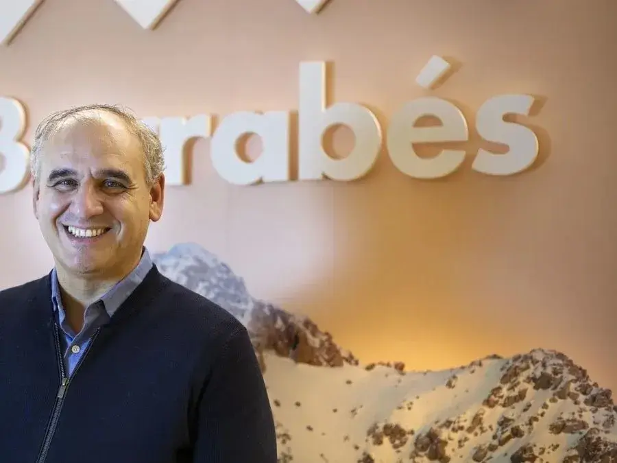 Carlos Barrabés sonríe ante el logo de Barrabés, con montañas nevadas de fondo.