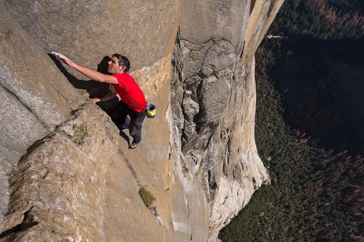Un escalador en solitario asciende una pared de roca imponente, capturando la esencia de un documental de escalada. La vista es vertiginosa, con un bosque denso abajo.