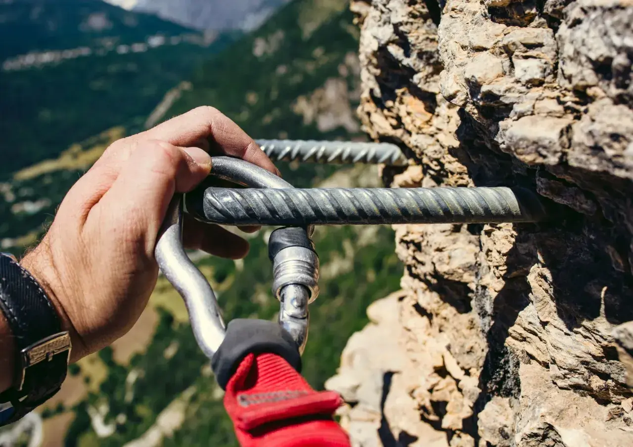 Mano enguantada se engancha a la **línea de vida vía ferrata** en una pared rocosa, con un paisaje montañoso al fondo.