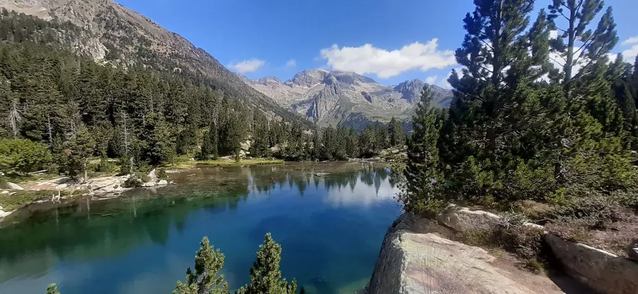 El ibonet de Batisielles, un espejo de agua turquesa rodeado de pinos y montañas imponentes, refleja el cielo azul y las nubes.