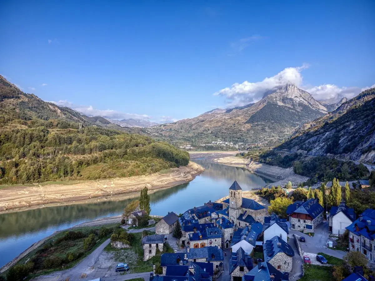 Pueblo de piedra junto a un embalse, con montañas nevadas de los picos de Huesca al fondo.