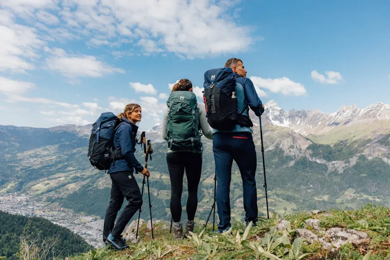 Tres excursionistas con mochilas y bastones admiran un paisaje montañoso. Sus prendas, ideales para distintos tipos de goretex, los protegen del clima.