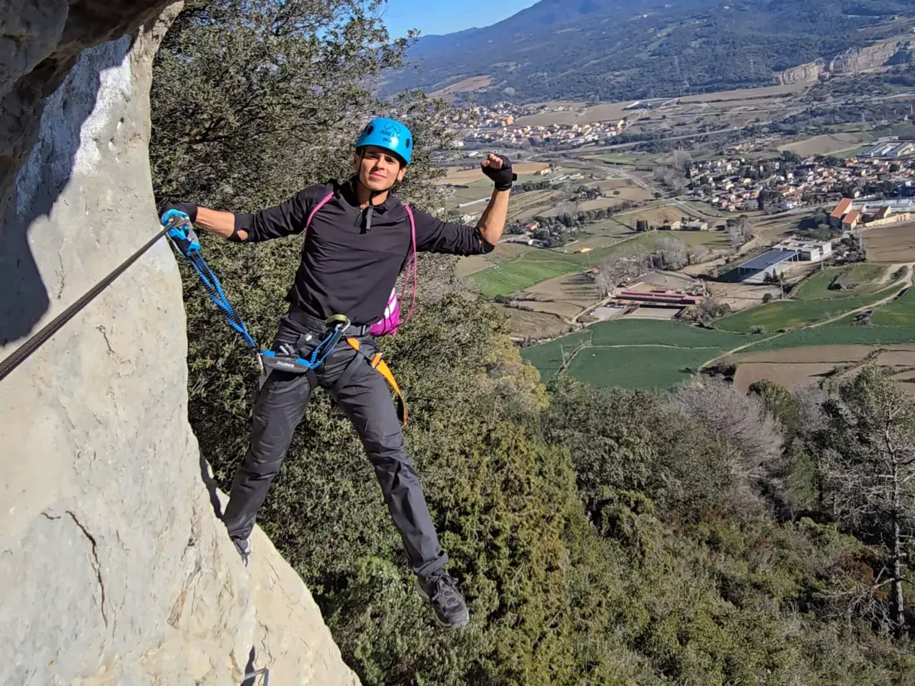 Un escalador sonriente, con casco azul, disfruta de la vía ferrata, un ejemplo de los diversos tipos de escalada, con un pueblo y campos verdes de fondo.