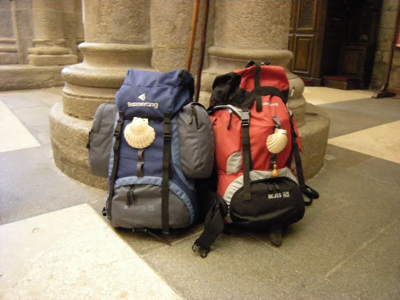 Dos mochilas, una azul y otra roja, con conchas de peregrino, listas para el camino. El tamaño de la mochila es clave para el Camino de Santiago.
