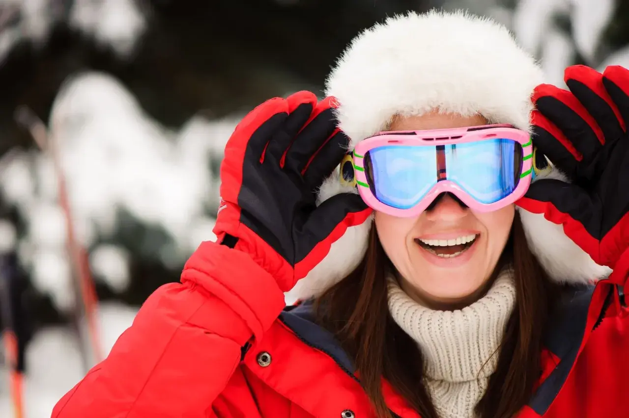 Mujer sonriente con gorro de piel y gafas de esquí rosas, lista para la nieve. Explora las categorías de gafas de esquí para encontrar tu estilo.