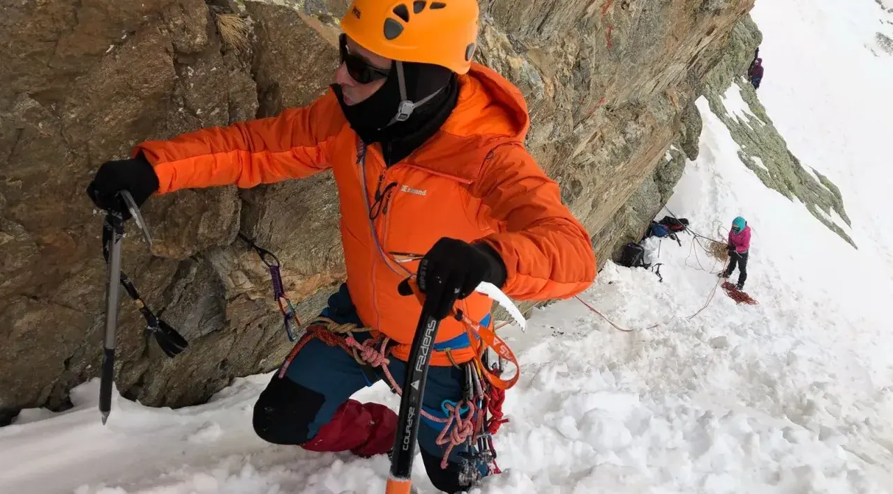 Alpinista con casco naranja y chaqueta brillante usa un piolet para escalar una pared rocosa nevada.