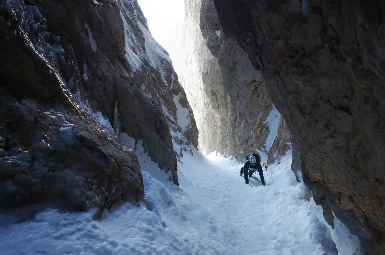 Alpinista asciende por un estrecho cañón nevado, usando piolet y crampones. La estaca de nieve se ve firme bajo sus pies.