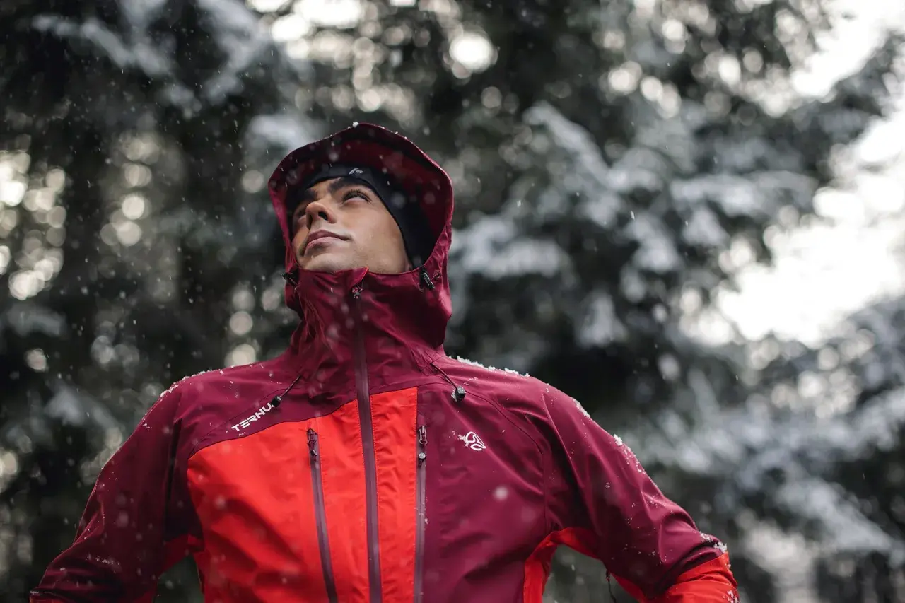 Hombre con ropa para ir a la montaña, con capucha y chaqueta impermeable, bajo una nevada.