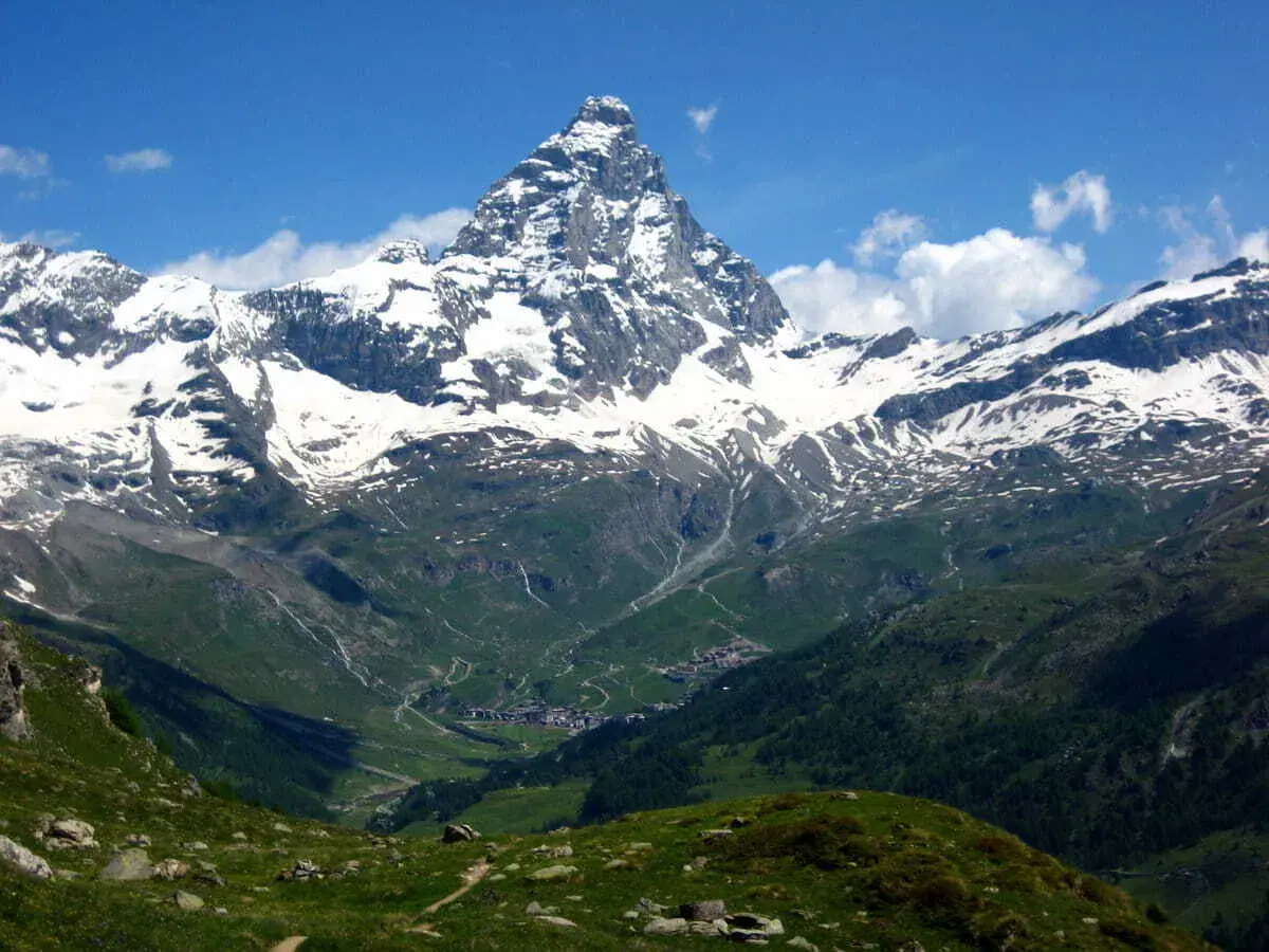 Majestuoso pico nevado se alza sobre un valle verde con un pueblo. El camino de senderismo gr5 etapes serpentea entre las colinas.